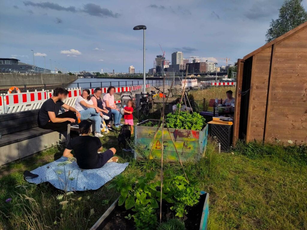 Eine Gruppe von Menschen vor dem Depot im Oberhafen Garten. Die Abendsonne scheint. Im HIntergrund sind die Elbbrücken und Gebäude zu sehen.