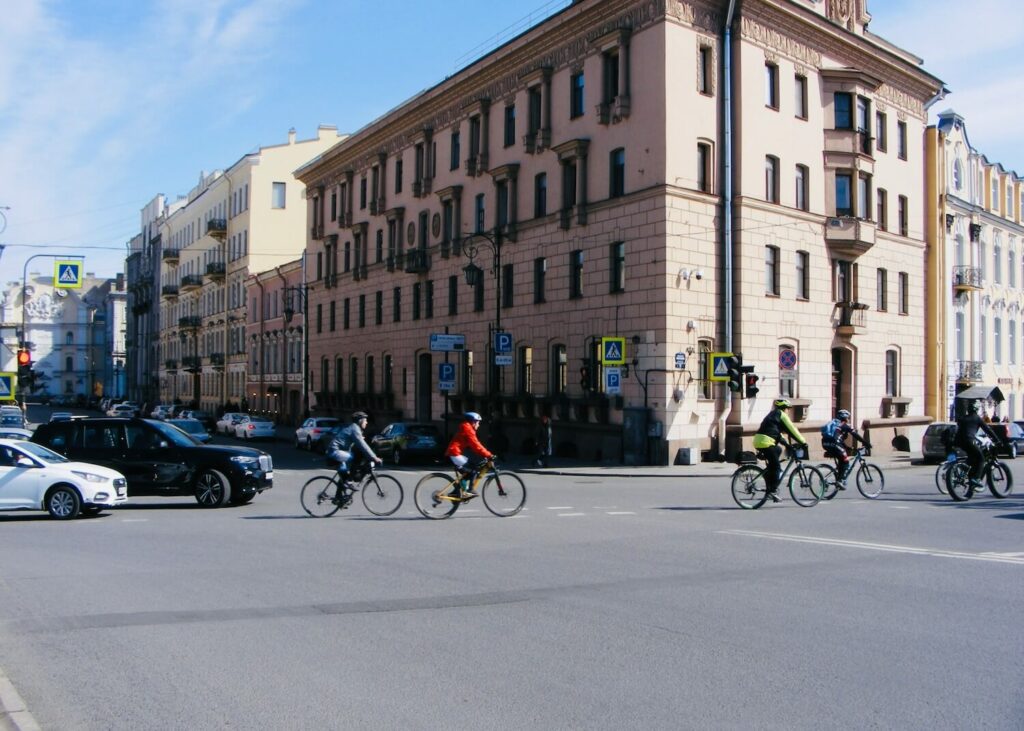 Eine Straßenkreuzung in der Stadt auf der Radfahrer·innen und Autos entlang fahren. Im Hintergrund stehen mehrere dreistöckige Mehrfamilienhäuser.